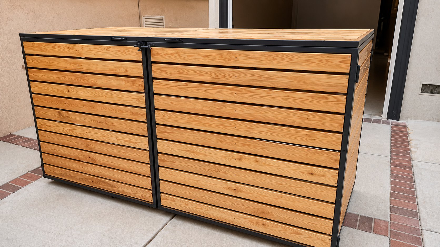 Cedar and steel trash can hide installed against home wall, brick-bordered concrete pad