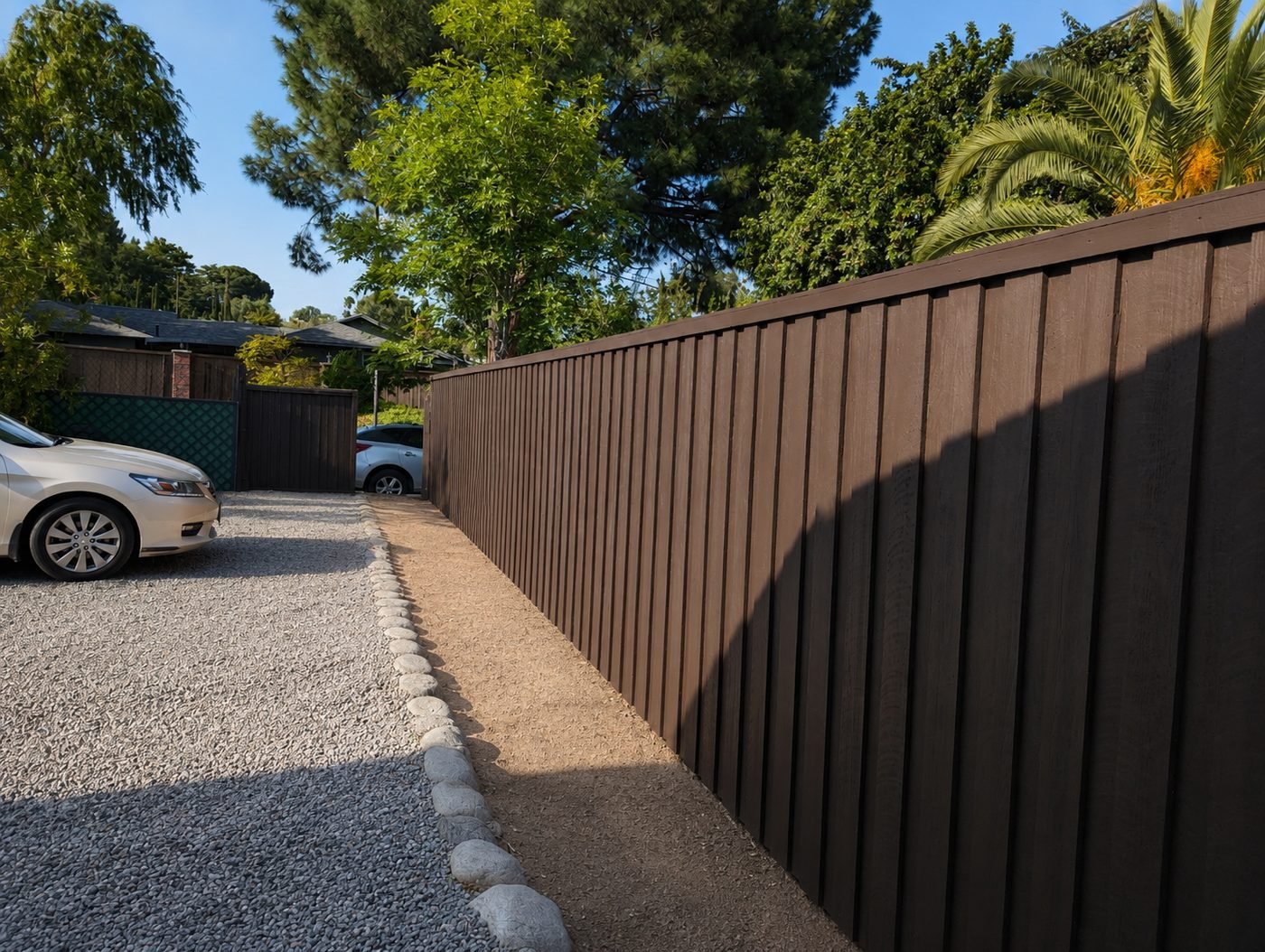 New board-and-batten cedar fence along a driveway with pea gravel and cobblestone edging — deep chocolate brown stain — ProHands Home Improvement Los Angeles