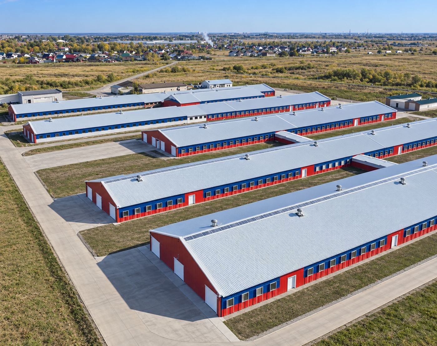 Aerial view of a long row of blue-and-red steel-framed livestock barns we built in Russia, set in open fields next to a village