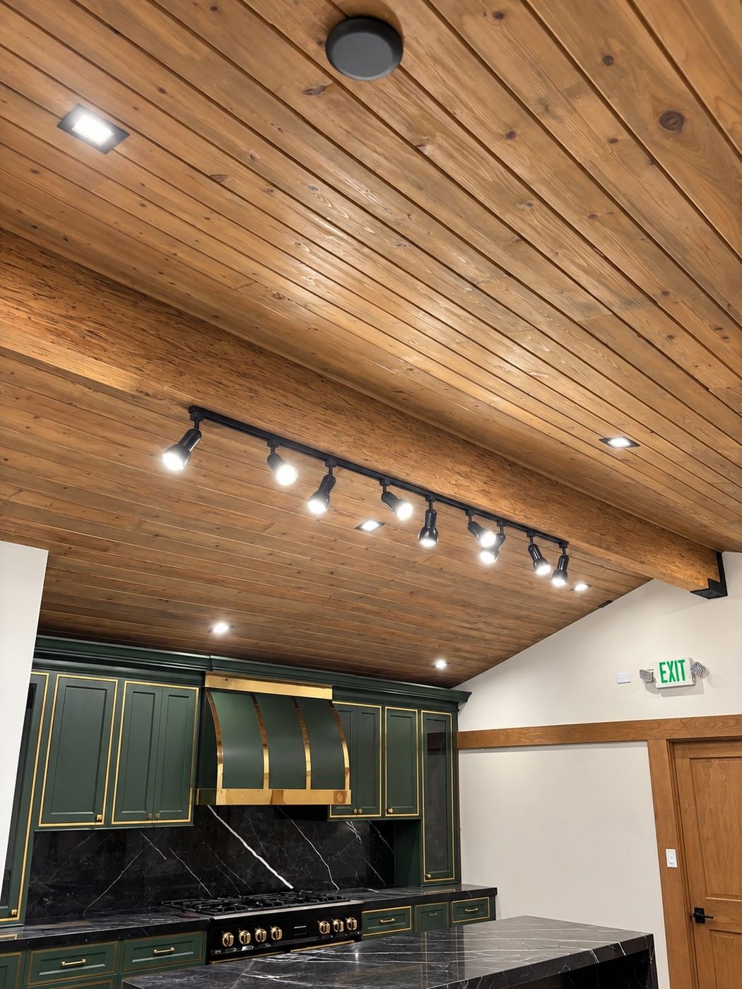 Display kitchen inside the finished showroom — hunter-green shaker cabinets with brushed-brass trim, brass range hood and black marble counters, set under the new knotty-pine shiplap ceiling and track lighting we installed — ProHands Los Angeles