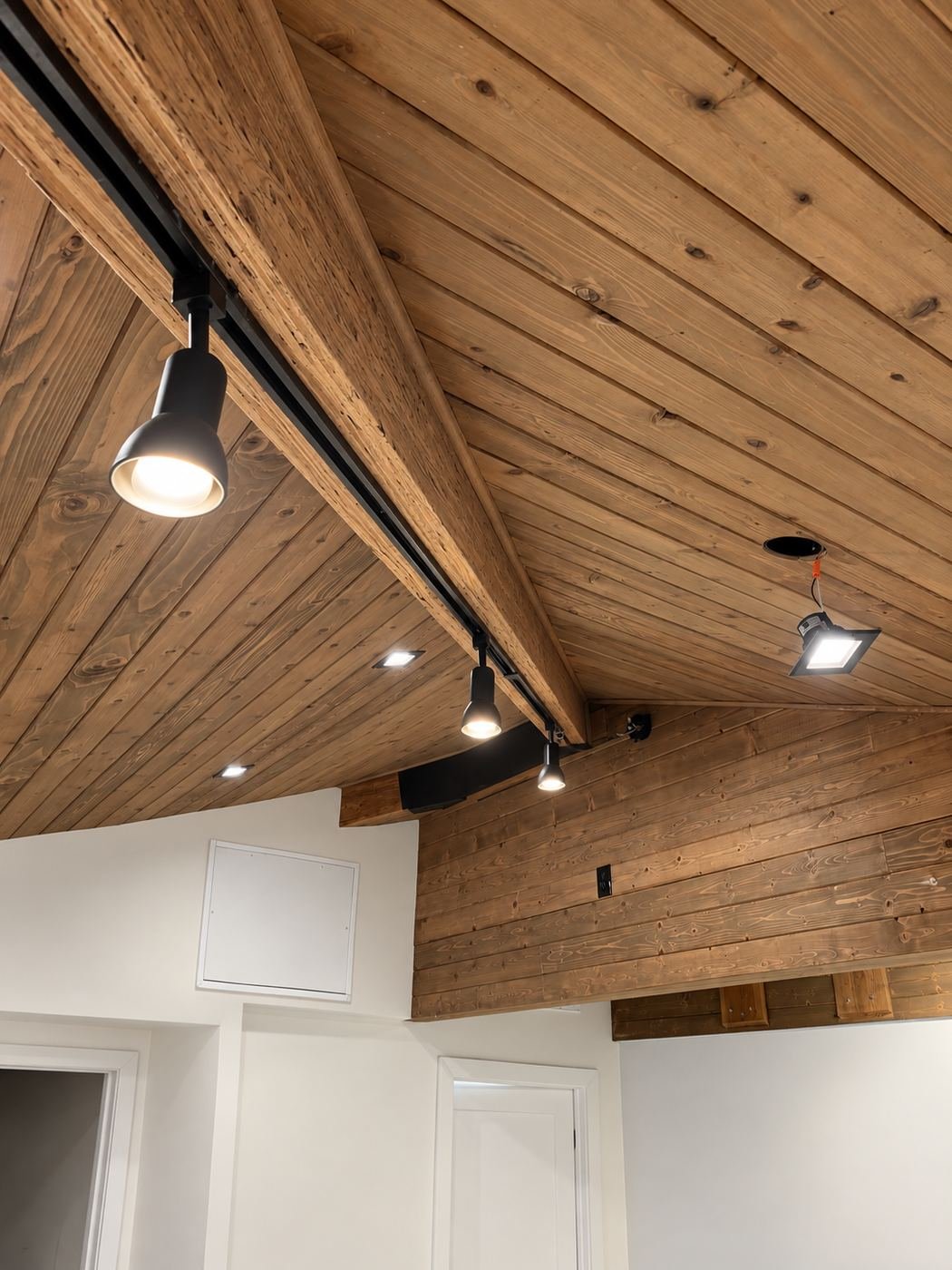 Detail of a black track-lighting rail bolted along an engineered-wood ridge beam against the new knotty-pine shiplap ceiling — ProHands Los Angeles kitchen showroom buildout