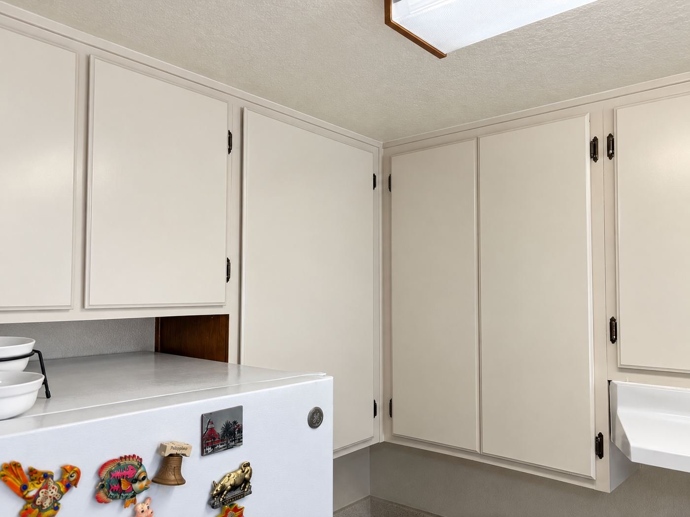 Wide view of refinished warm-white kitchen uppers and corner cabinets next to a white fridge, ceiling fluorescent light with wood trim, Glendale — ProHands