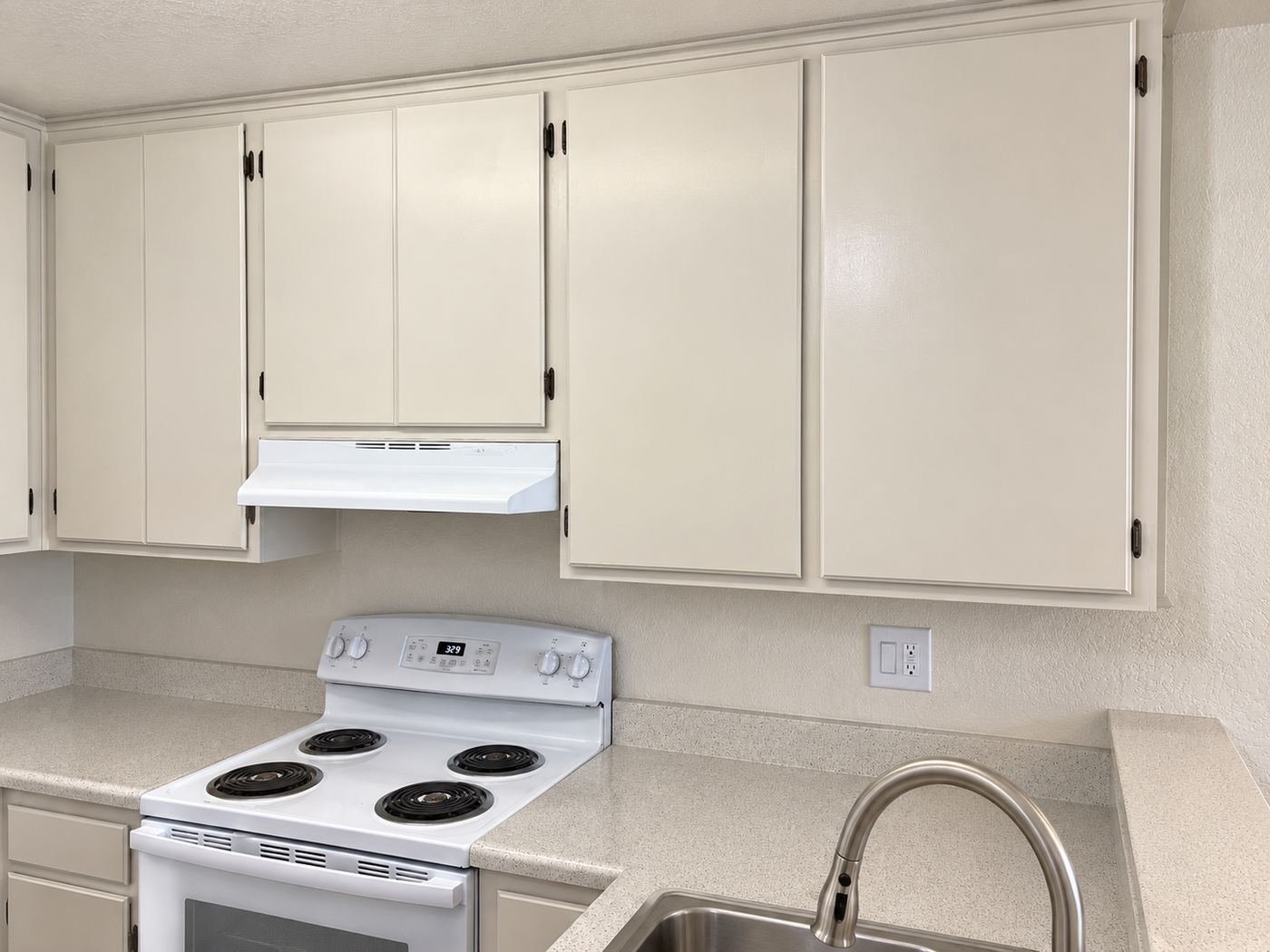 Wide view of a Glendale kitchen after cabinet refinish — warm-white uppers and lowers, white electric range, stainless sink, matching light speckled quartz counters — ProHands