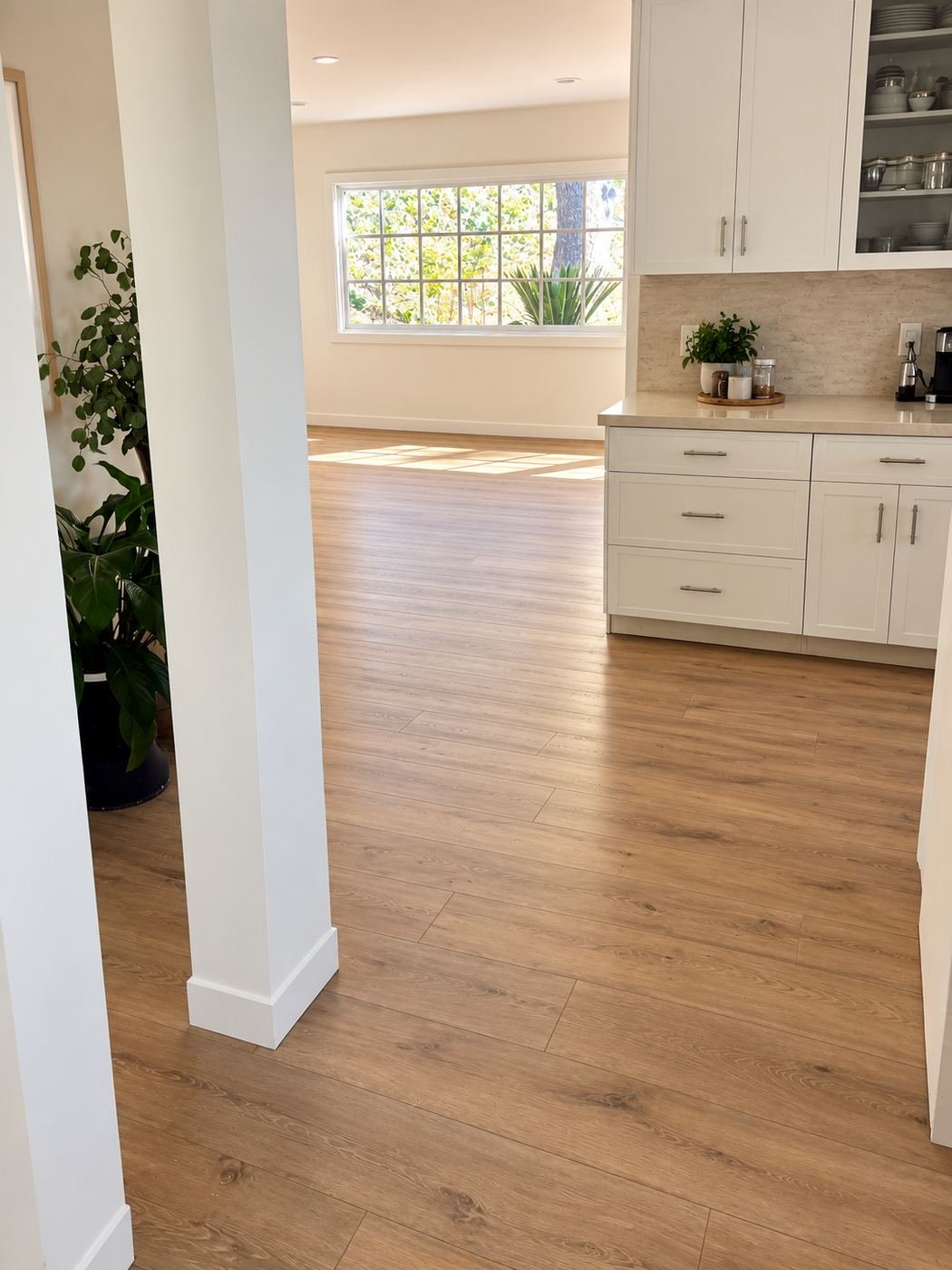 Hallway to kitchen transition — same warm-oak LVP floor runs through the doorway in one unbroken field, white shaker cabinets with quartz counter and tile backsplash on the right — ProHands Los Angeles