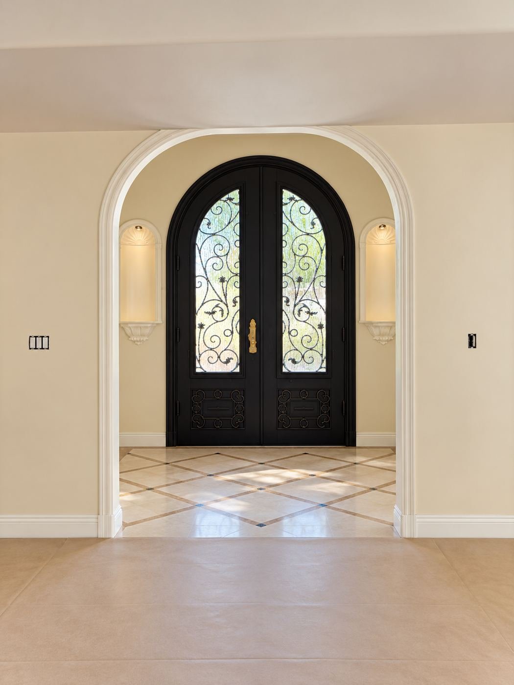 Formal reception area looking through a large interior arch toward the mansion's matching black iron entry doors with scrolled grilles — picture-frame moldings on every wall, travertine floor — ProHands LA mansion remodel