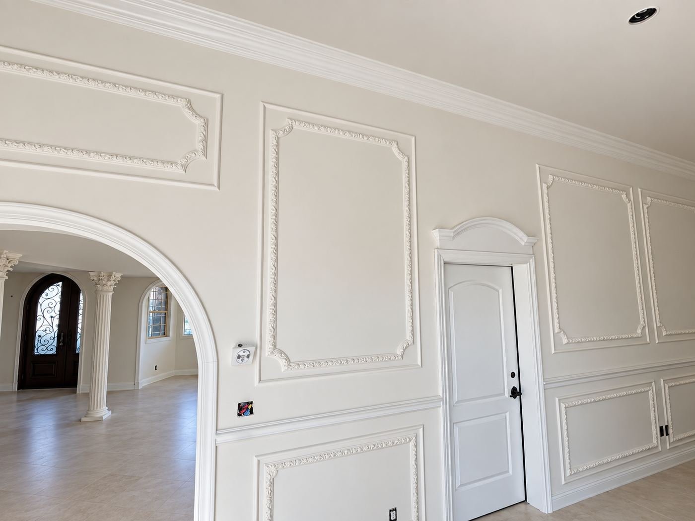 Transitional formal hall of a Mediterranean mansion with elaborate picture-frame wall moldings, a column visible through the arch and a view toward the iron entry doors — ProHands Los Angeles