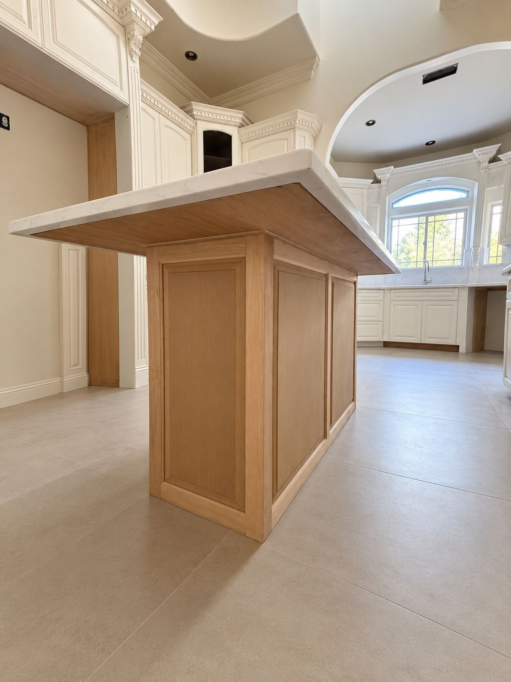 Side view of the second kitchen island — hand-wrapped wood base in warm oak with carved brackets, polished marble countertop with ogee edge, large travertine tile floor — ProHands mansion remodel LA