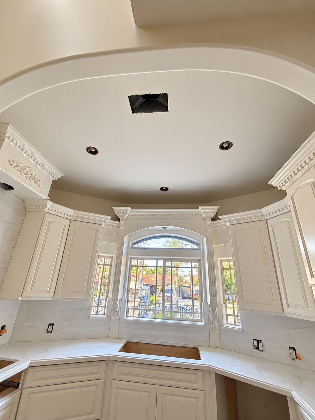 Second kitchen area with a curved ceiling detail, bank of white raised-panel upper cabinets, cream crown molding and a large arched window above a quartz counter — ProHands mansion remodel LA