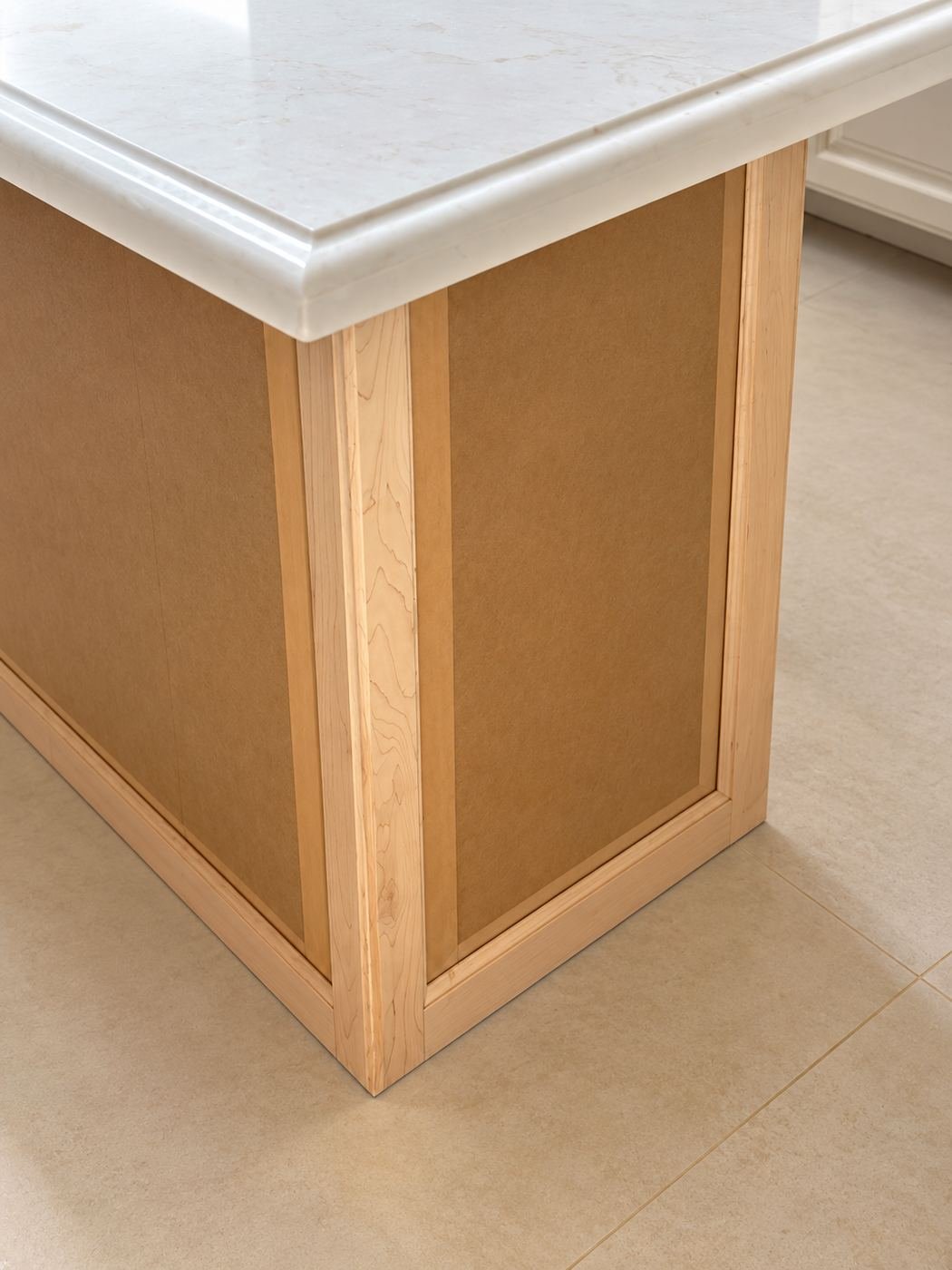 Close-up of the wood-wrapped kitchen island base — warm honey oak panels with picture-frame molding, picture-frame ogee marble top edge — ProHands mansion remodel LA
