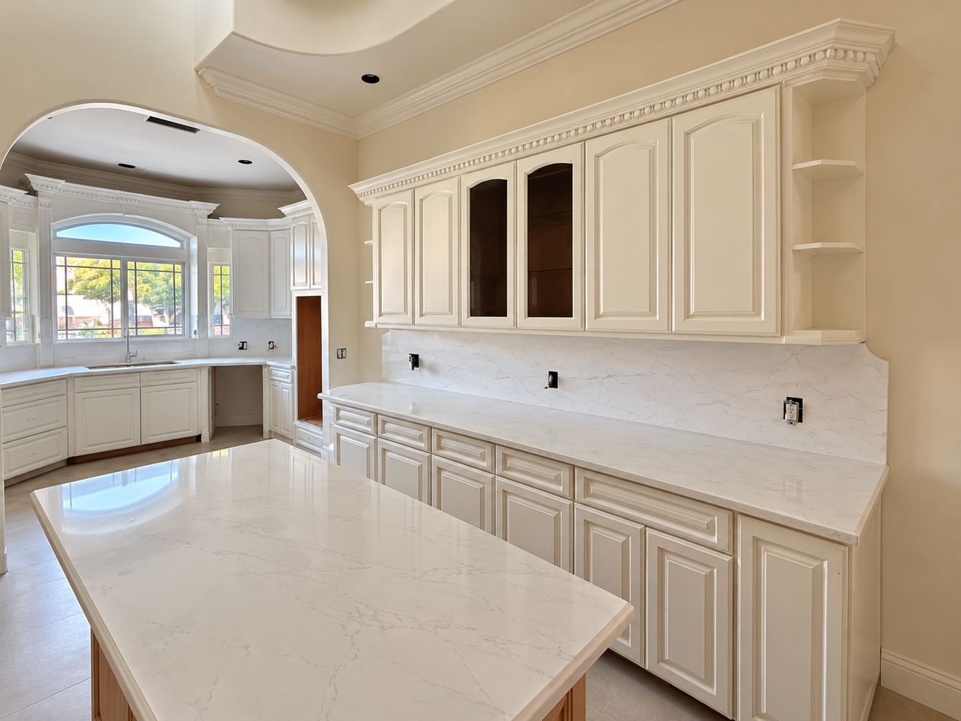 Large arched window above the kitchen counter — marble countertops, cream raised-panel cabinets with carved brackets below, view out to mature trees — ProHands mansion remodel LA
