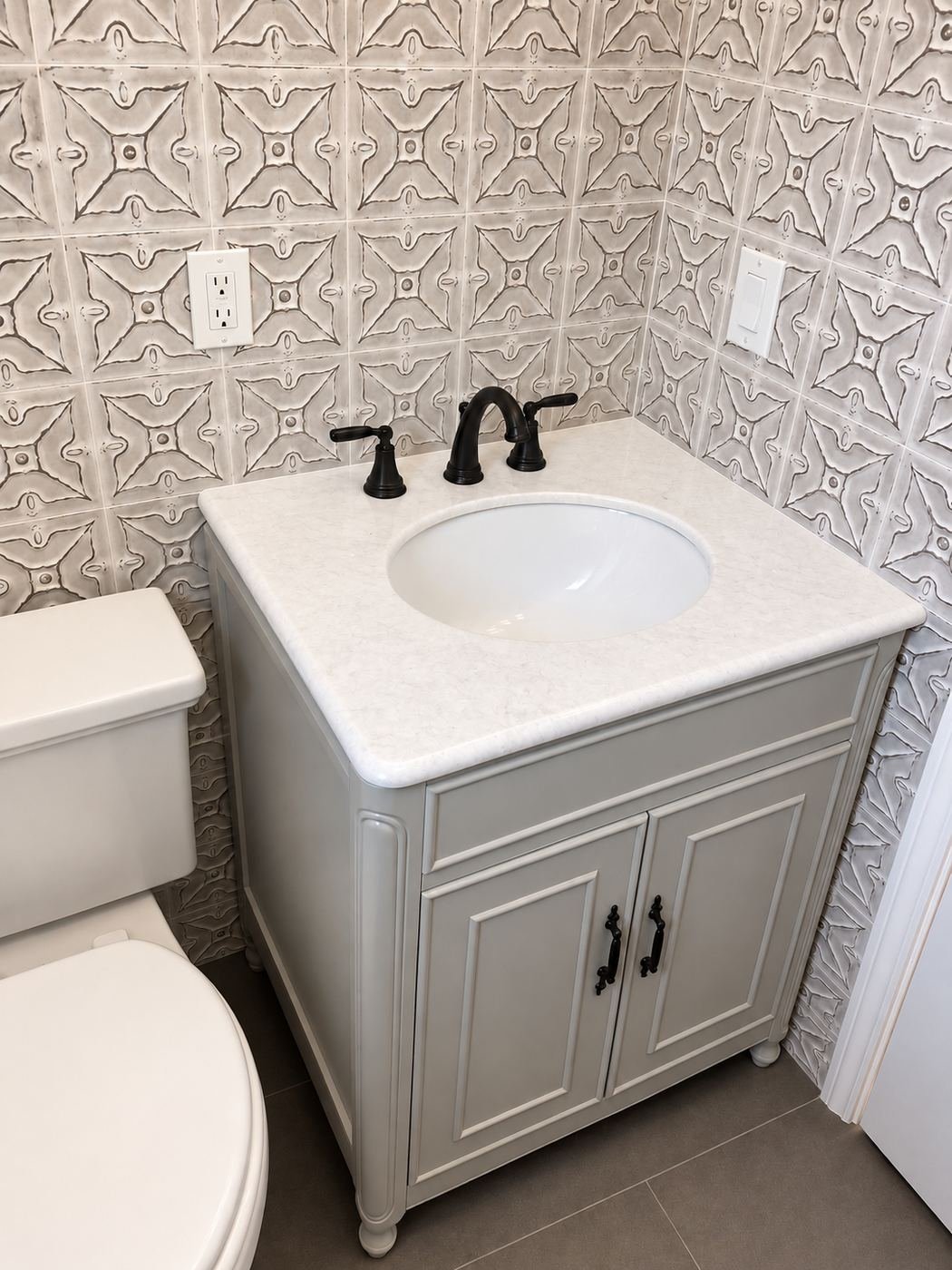 Powder-room vanity under a tin-tile wall — carved cream vanity with marble top, oval sink, black bridge-style faucet and matching black knob hardware — Mediterranean mansion remodel, Los Angeles