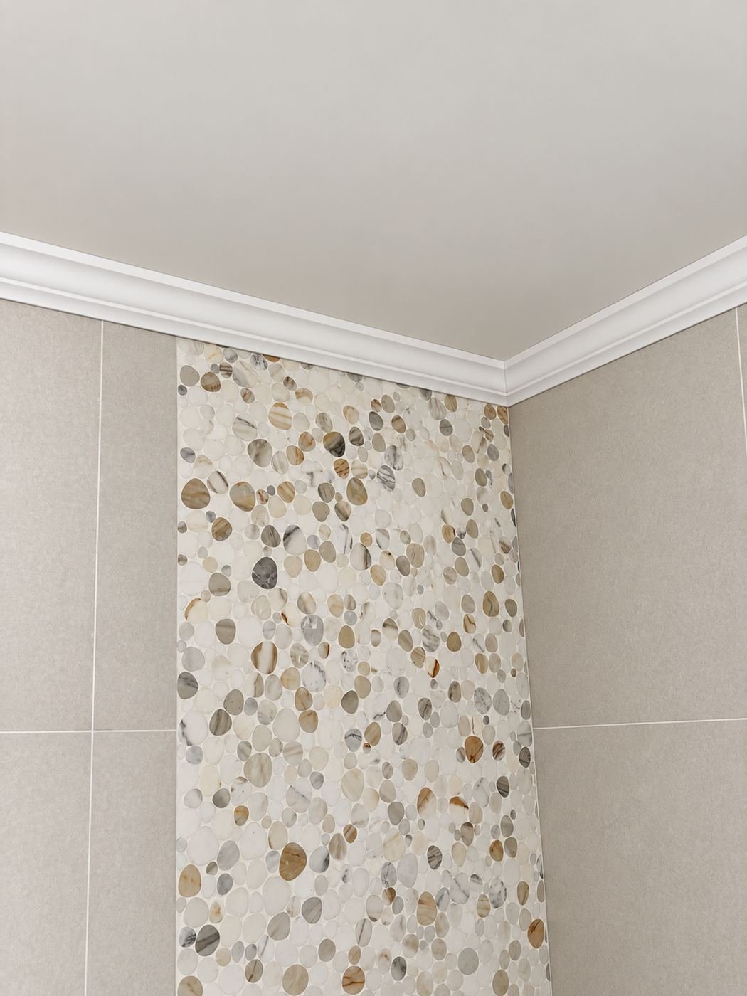 Pebble-tile vertical strip accent on a bath wall — matte beige background tile with a wide river-pebble inlay running floor to ceiling, warm-cream crown molding above — ProHands LA mansion remodel