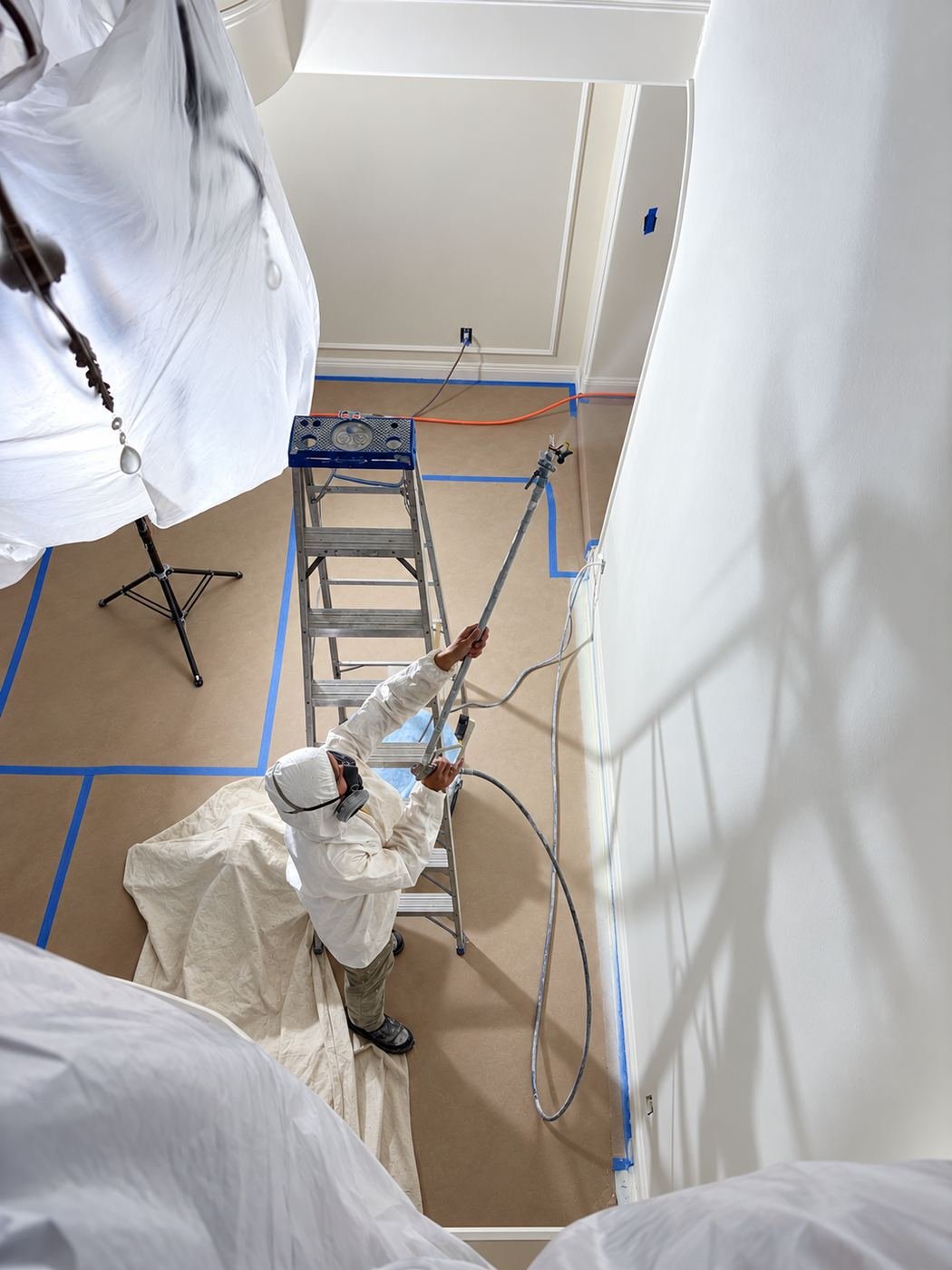 Tyvek painter on ladder with airless under plastic-wrapped chandelier — ProHands LA mansion