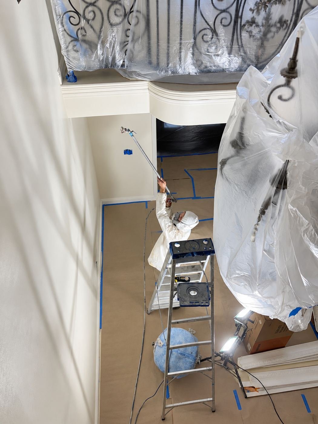 ProHands painter on a step-ladder with an airless spray gun, Tyvek suit and respirator, working inside a large arched opening framed by white moldings — wrought-iron rail from Chapter 1 visible — Mediterranean mansion remodel, LA