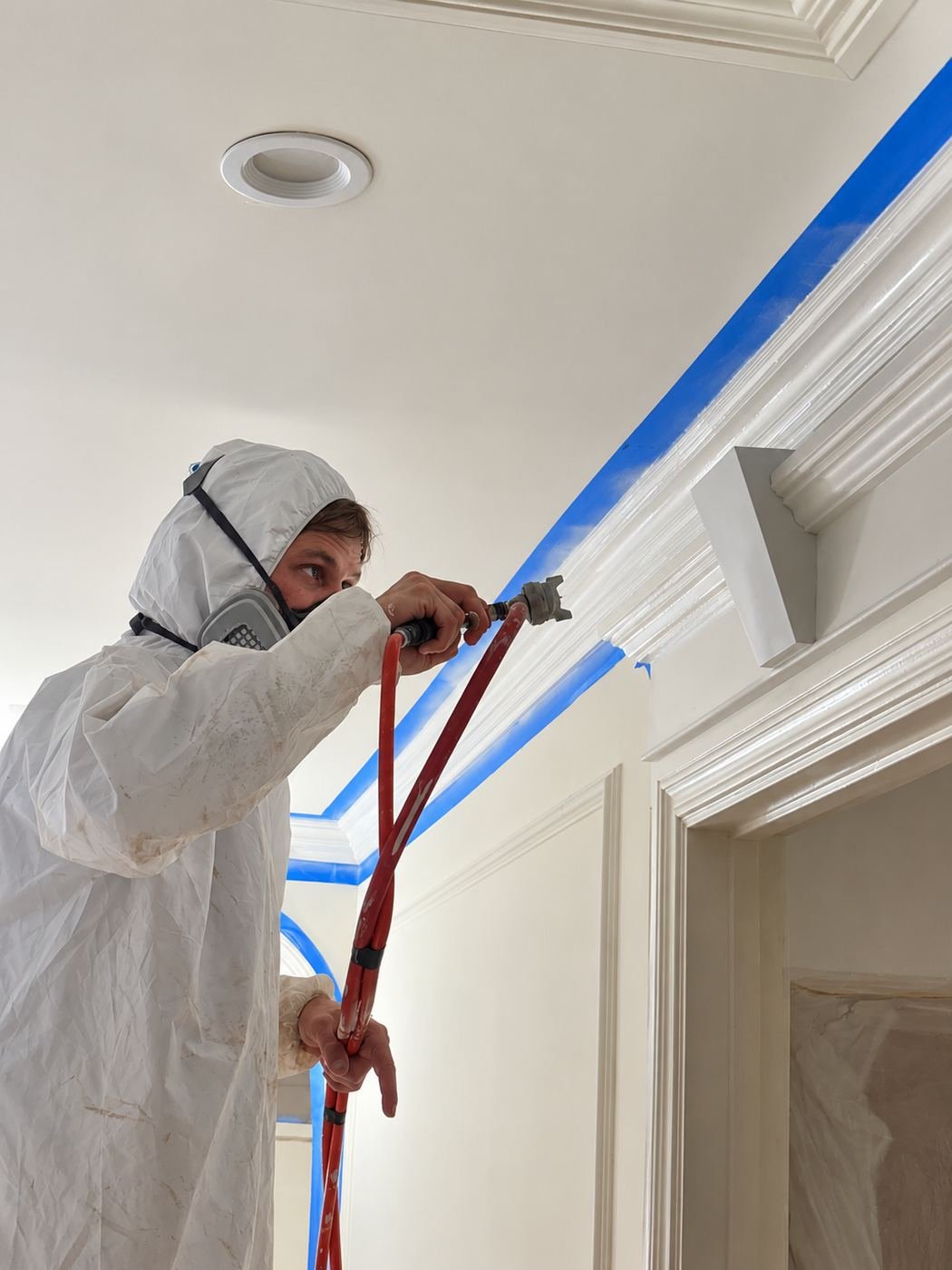 Close-up of a ProHands painter in Tyvek suit and respirator airless-spraying along a crown-molding line taped off with blue painter's tape — Mediterranean mansion remodel, Los Angeles