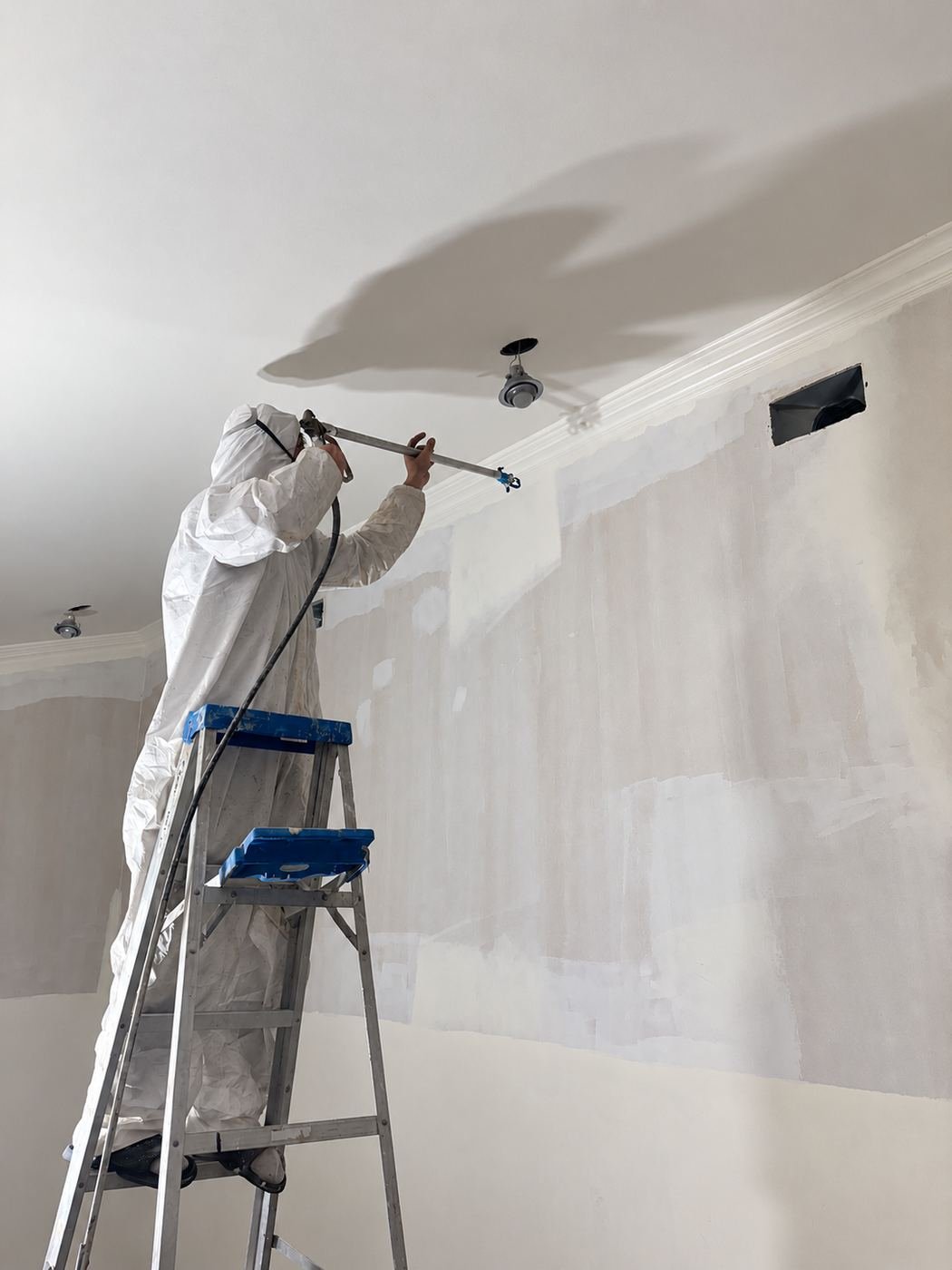 ProHands painter in Tyvek suit and respirator on a step-ladder airless-spraying the top of a wall corner, floor fully masked below — Mediterranean mansion remodel, Los Angeles