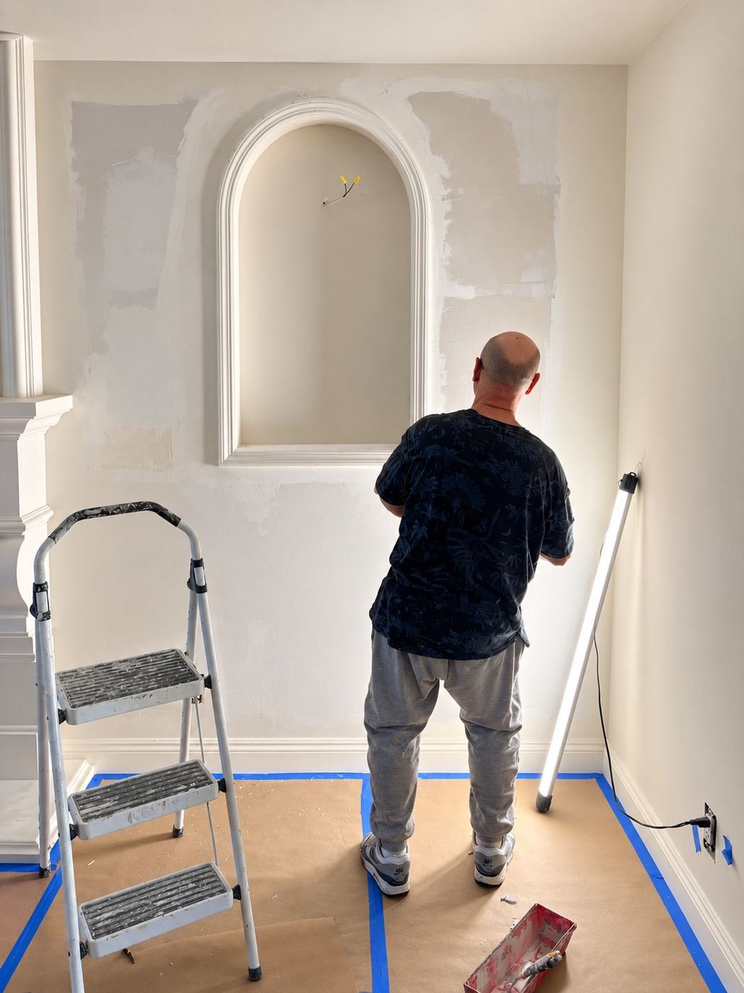 Worker on a two-step ladder brush-painting the inside curve of a large arched plaster niche, rosin paper and blue tape on the floor around the ladder — Mediterranean mansion remodel, Los Angeles