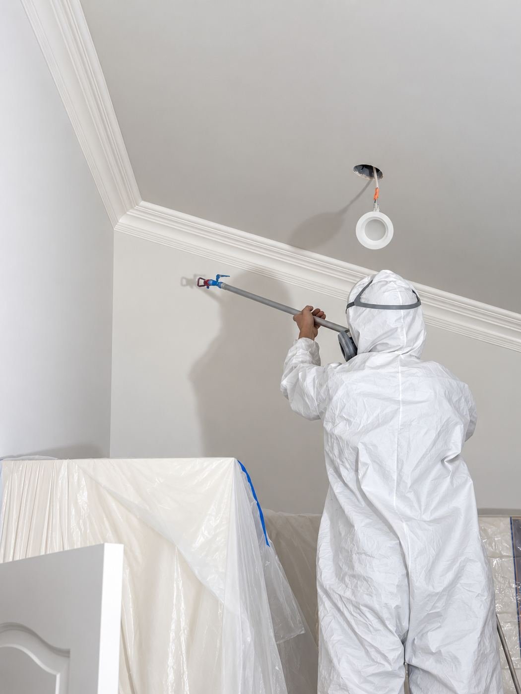 Painter in Tyvek suit using a roller on an extension pole to apply a ceiling patch coat, with the airless cart and plastic sheeting around the perimeter — Mediterranean mansion remodel, Los Angeles