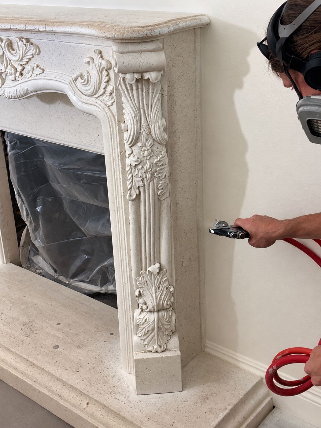 Close-up of a worker in a respirator finishing the carved side pilaster of an ornate limestone fireplace mantel with a small spray gun — plastic sheeting protecting the firebox opening — Mediterranean mansion remodel, Los Angeles