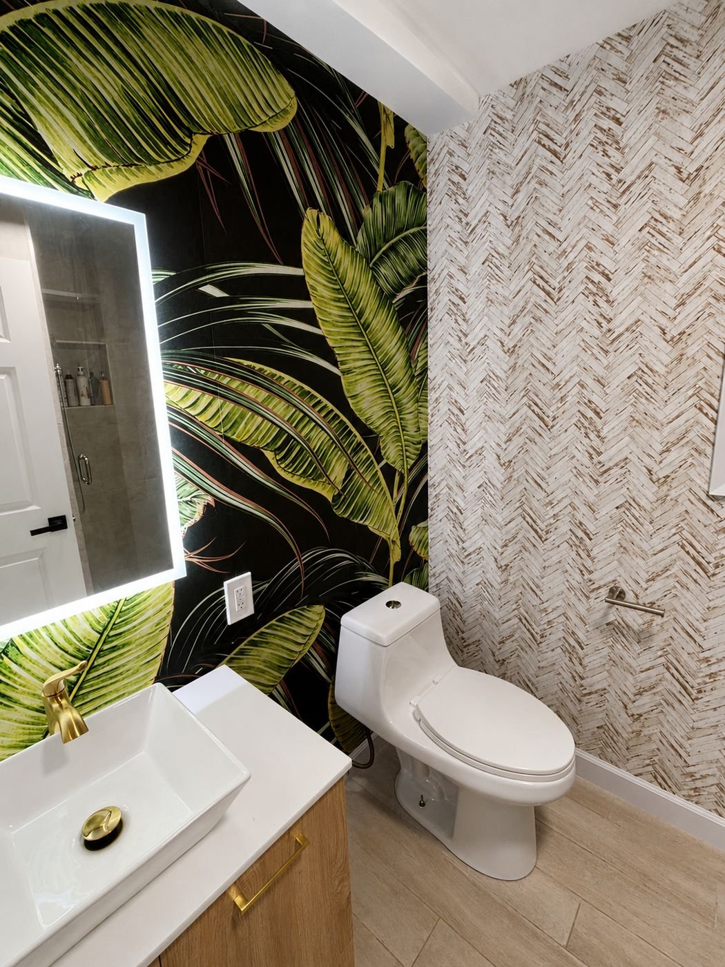 Powder room remodel in Los Angeles — dark banana-leaf tropical wallpaper, herringbone wood-look accent wall, light wood vanity with white vessel sink and gold hardware, backlit LED mirror, one-piece toilet — ProHands