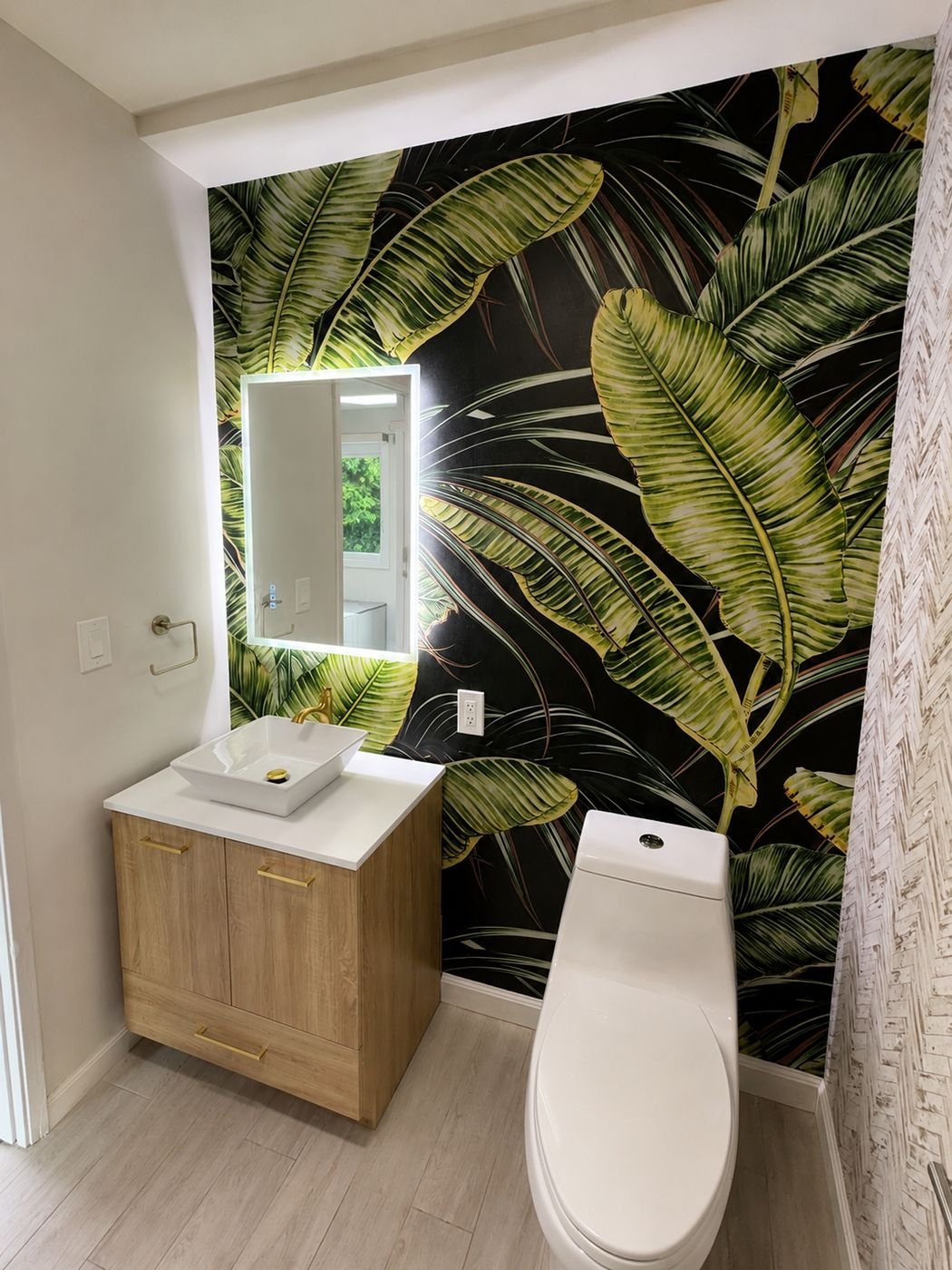 Modern powder room vanity with white vessel sink on light wood cabinet, gold faucet and pulls, LED backlit mirror with hidden wiring in the wall, banana-leaf wallpaper behind — ProHands Los Angeles
