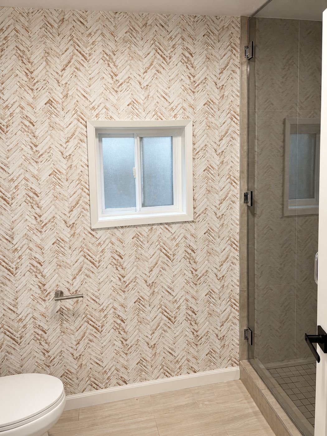Herringbone wood-look wallpaper accent wall with white-framed obscure-glass window and frameless glass shower door with grey tile shower niche — powder room remodel ProHands Los Angeles