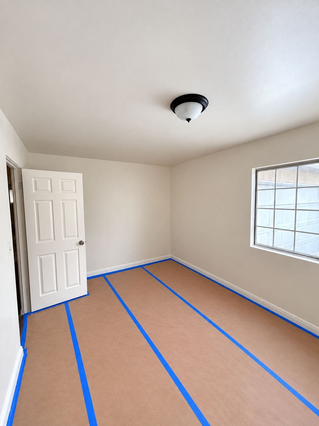 Bedroom prepped for paint during a Van Nuys interior restoration — client's laminate floor fully masked with rosin paper and blue tape against the baseboards, door and ceiling light left on