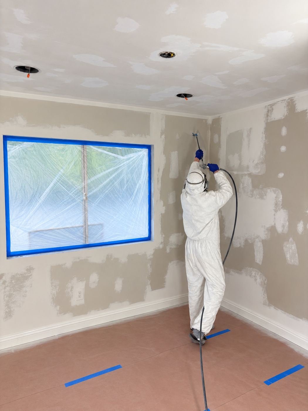 ProHands painter in full Tyvek suit and P100 respirator airless-spraying primer onto a wall full of drywall patches during the Van Nuys water-damage restoration — fresh new drywall after mold-affected sections were replaced