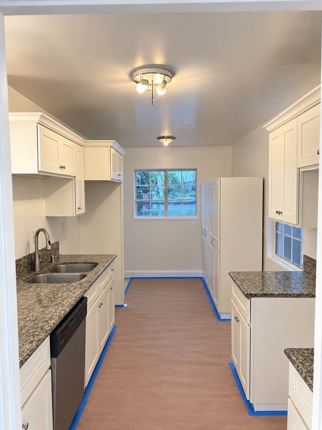 Finished galley kitchen inside the Van Nuys restoration — repainted ceiling and walls, existing white shaker cabinets with granite counters cleaned and left in place, client's laminate floor masked for final paint pass