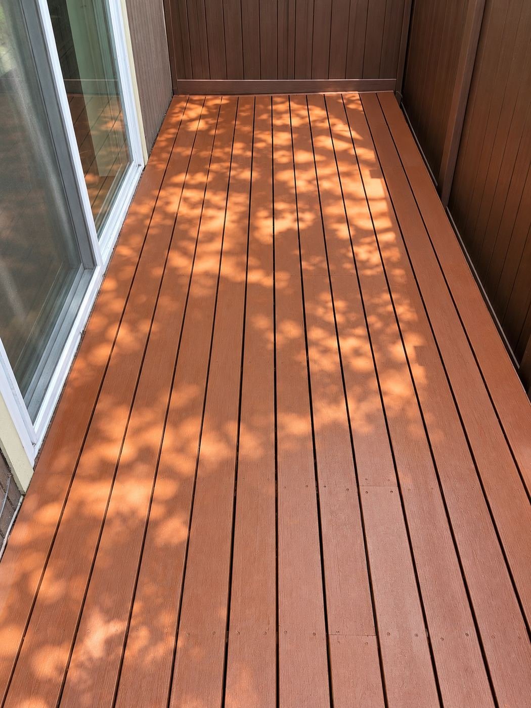 Restored wood deck in a warm cedar/mahogany stain with dappled sunlight from an overhead tree, matching stained siding and a white vinyl slider door — ProHands Los Angeles deck restoration