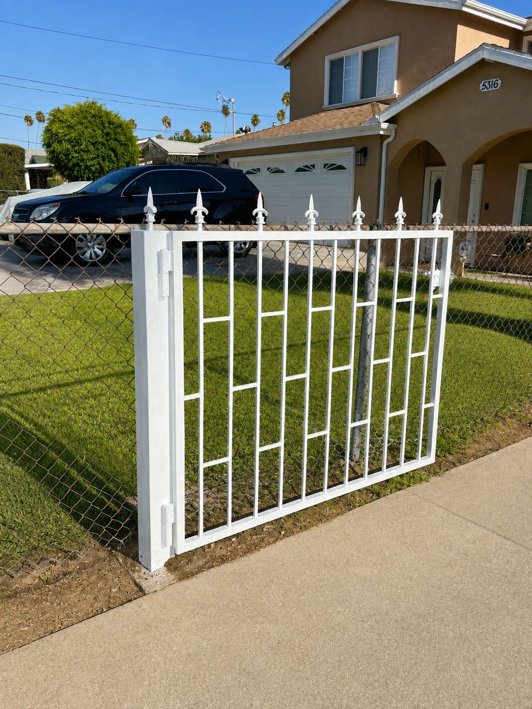 Freshly repainted white wrought-iron pedestrian gate with fleur-de-lis spikes, installed in front of an existing chain-link fence at a two-story stucco home in Los Angeles — ProHands